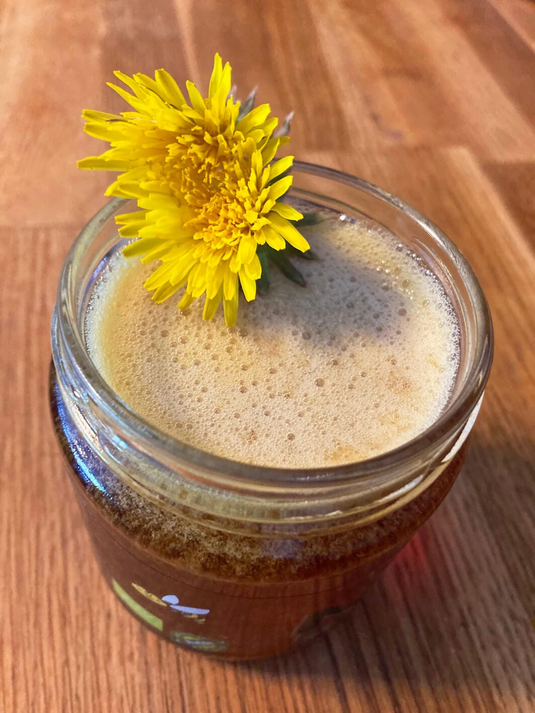 Homemade dandelion syrup in a glass jar, topped with a fresh dandelion flower, sitting on a wooden table.